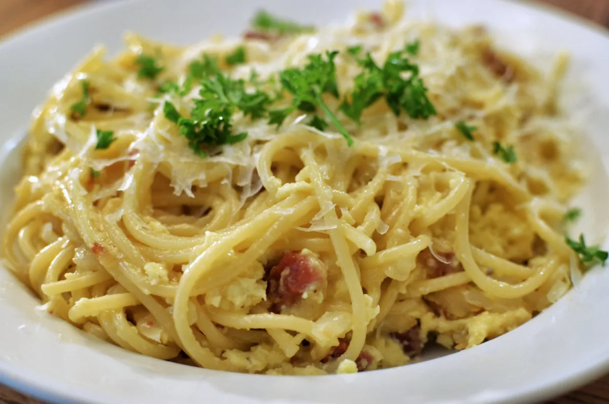 Close-up of spaghetti carbonara topped with shaved Parmesan, chopped parsley and pieces of pancetta in a white bowl