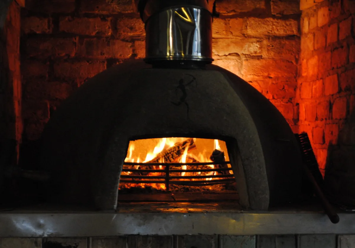 A dome-shaped wood-fired oven with a log fire burning inside, set against a red brick wall