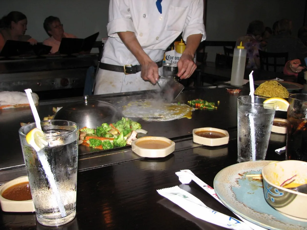 A hibachi chef in a white jacket cooking broccoli, carrots and fried rice on a steaming teppanyaki grill, with diners' drinks and sauce cups on the table edge